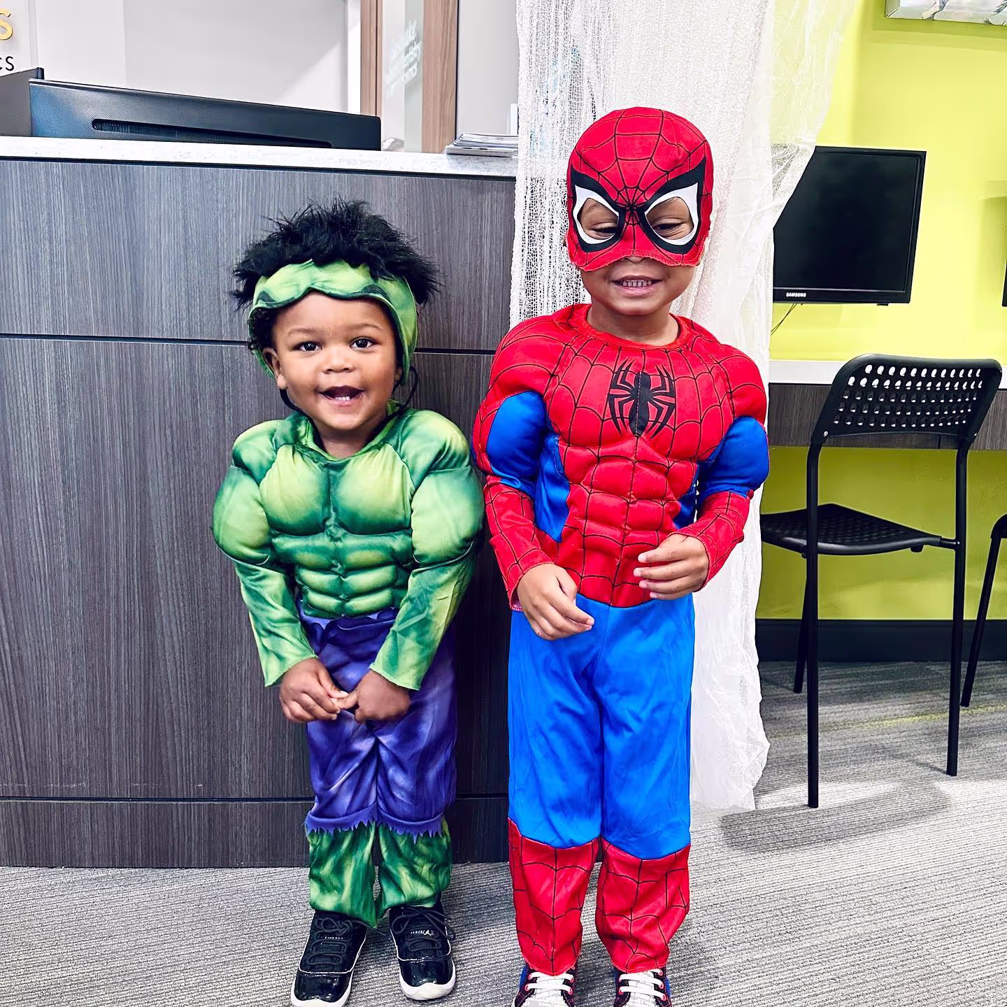 Two young children standing indoors dressed in superhero costumes, one as the Hulk and the other as Spider-Man.