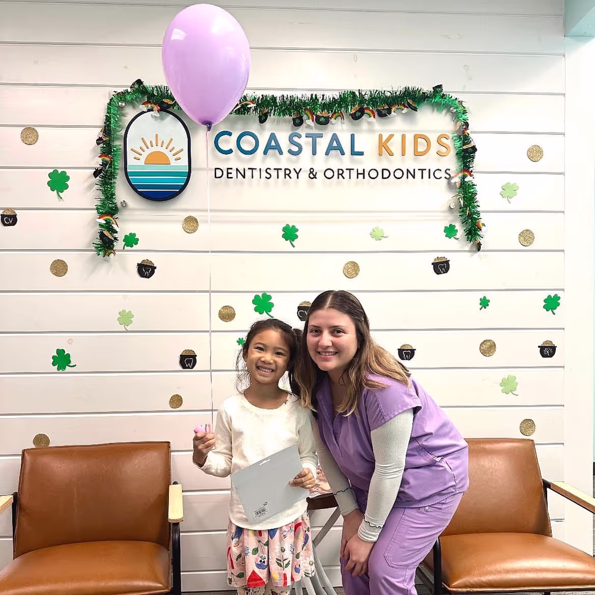 Smiling female dentist in purple scrubs poses with a young girl holding a balloon and a paper in a dental office decorated with shamrocks.