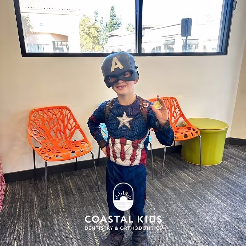 Young child dressed in a Captain America costume holding a gold coin, standing in a modern waiting room with orange chairs and a green ottoman.