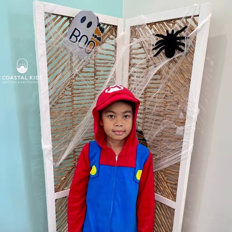 Child dressed in a Mario costume standing in front of a Halloween-themed screen with spider webs, a black spider, and a ghost decoration that says 'Boo'.