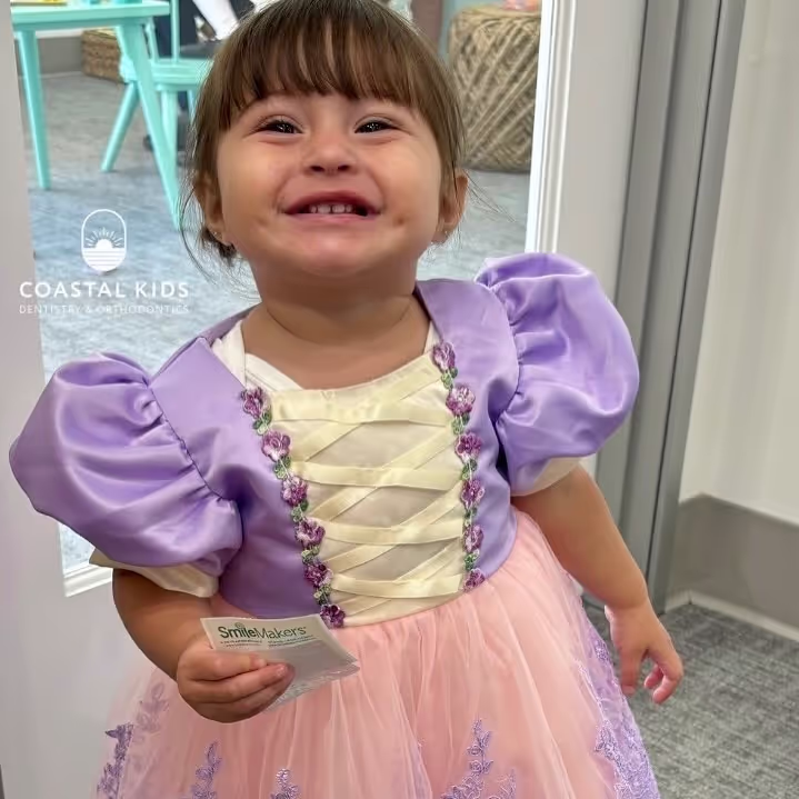 Young girl smiling widely, wearing a purple and pink princess dress, holding a SmileMakers card indoors.