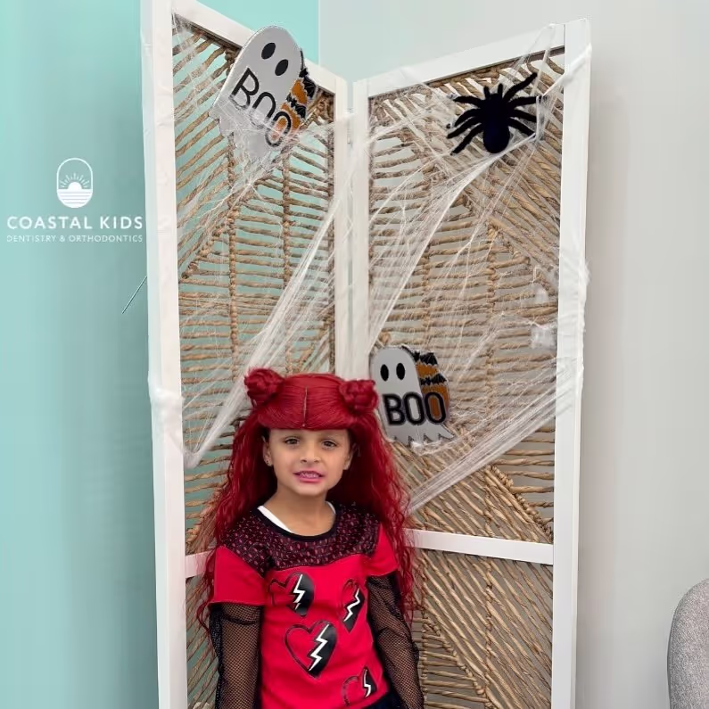 Young girl with long red hair styled in two buns wearing a red and black outfit standing in front of a Halloween-themed screen with ghost and spider decorations.