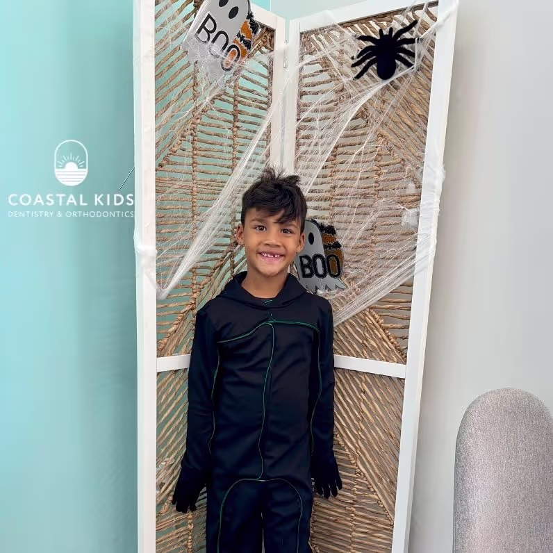 Smiling boy in a black outfit standing in front of a Halloween-decorated screen with spider webs, a black spider, and ghost signs saying 'BOO'.