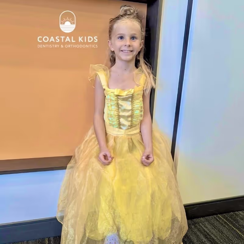 Smiling young girl wearing a yellow princess dress standing indoors in front of a Coastal Kids Dentistry & Orthodontics sign.