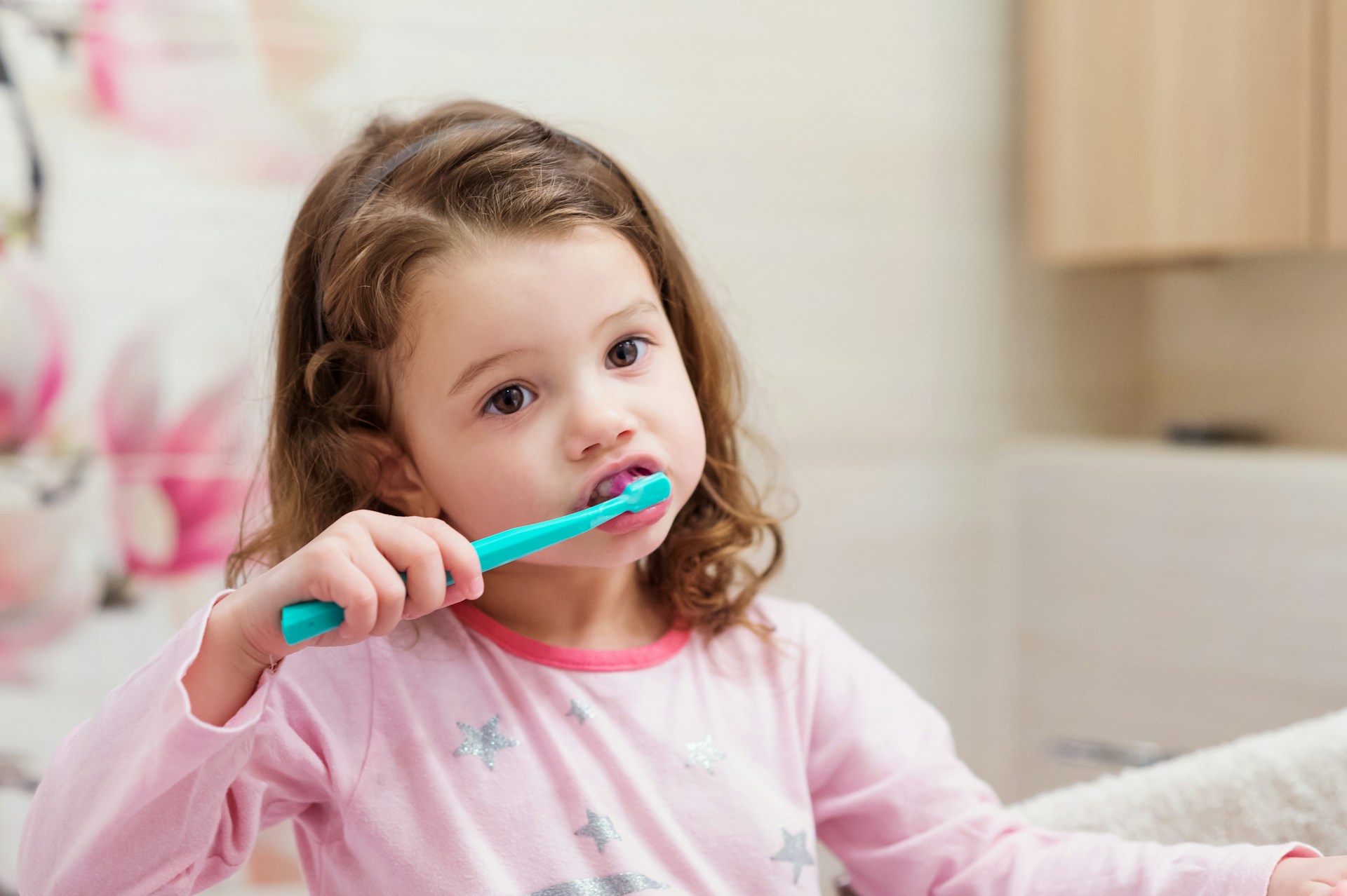 Young girl with curly brown hair brushing her teeth with a turquoise toothbrush in a bathroom.