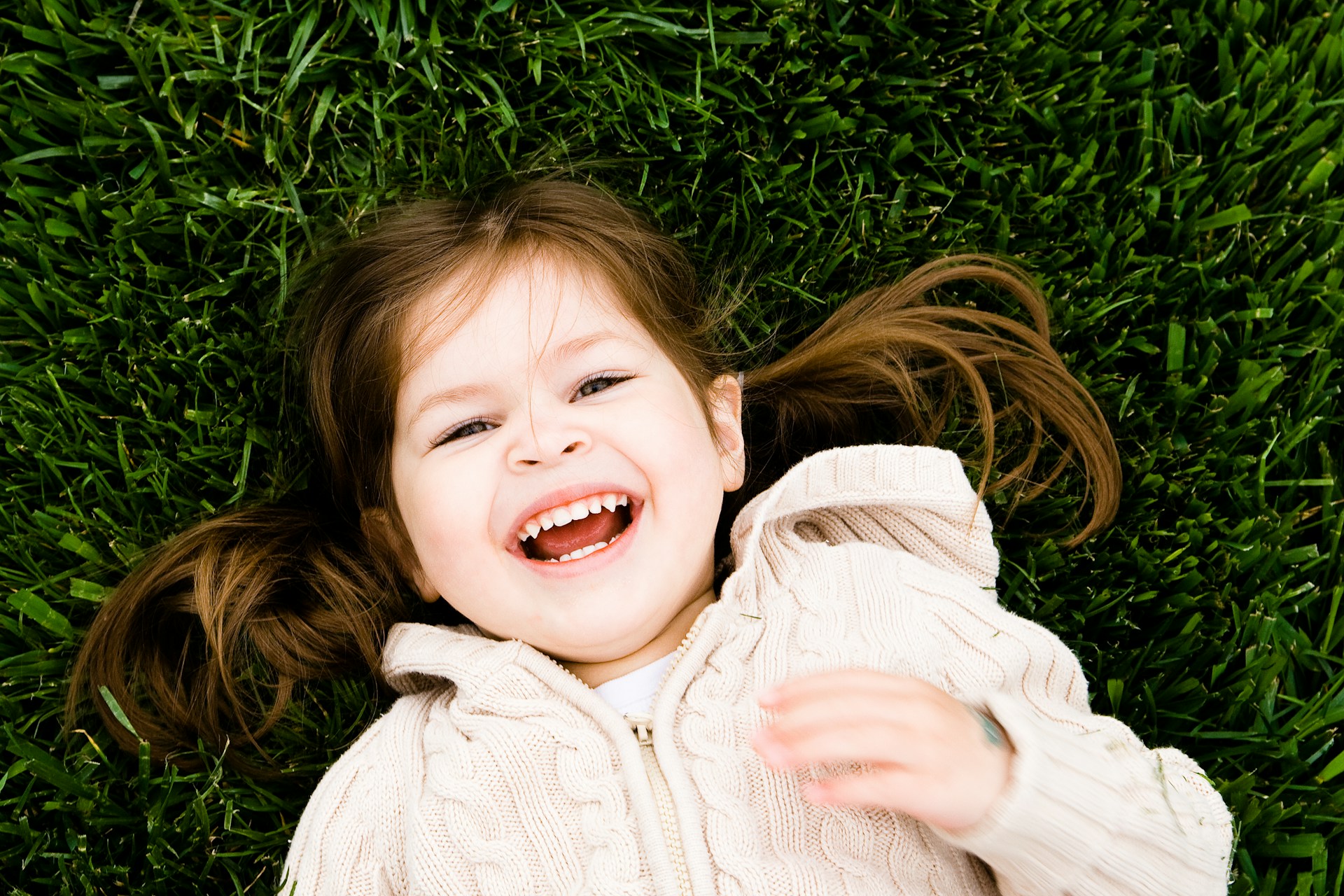 Smiling young girl with brown hair lying on green grass wearing a cream-colored knitted sweater.