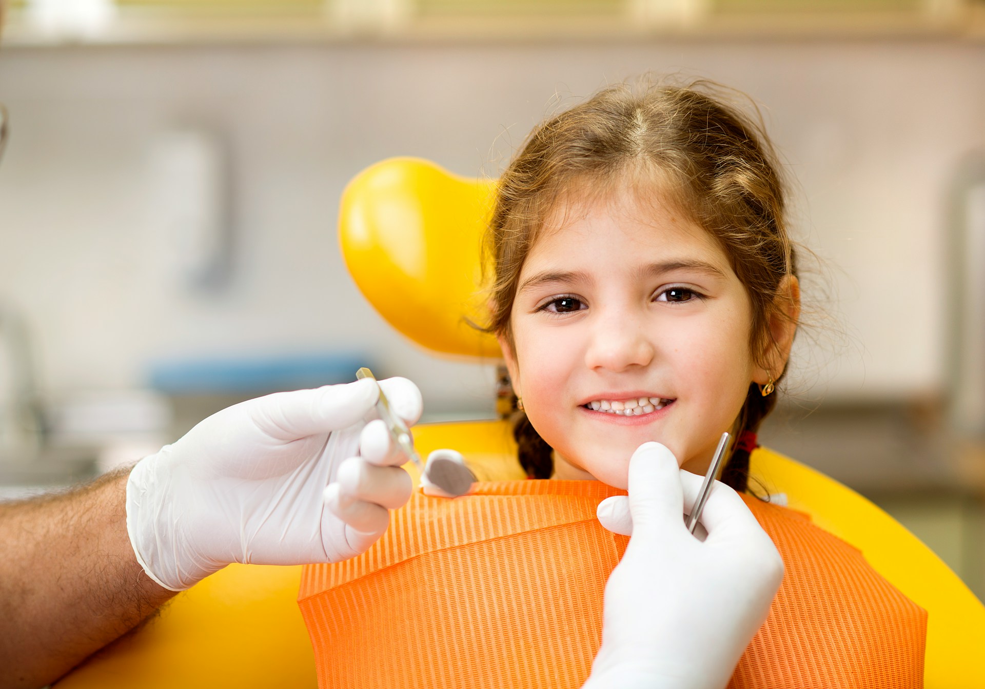 Smiling young girl sitting in a dental chair with dentist's gloved hands holding dental tools near her face.