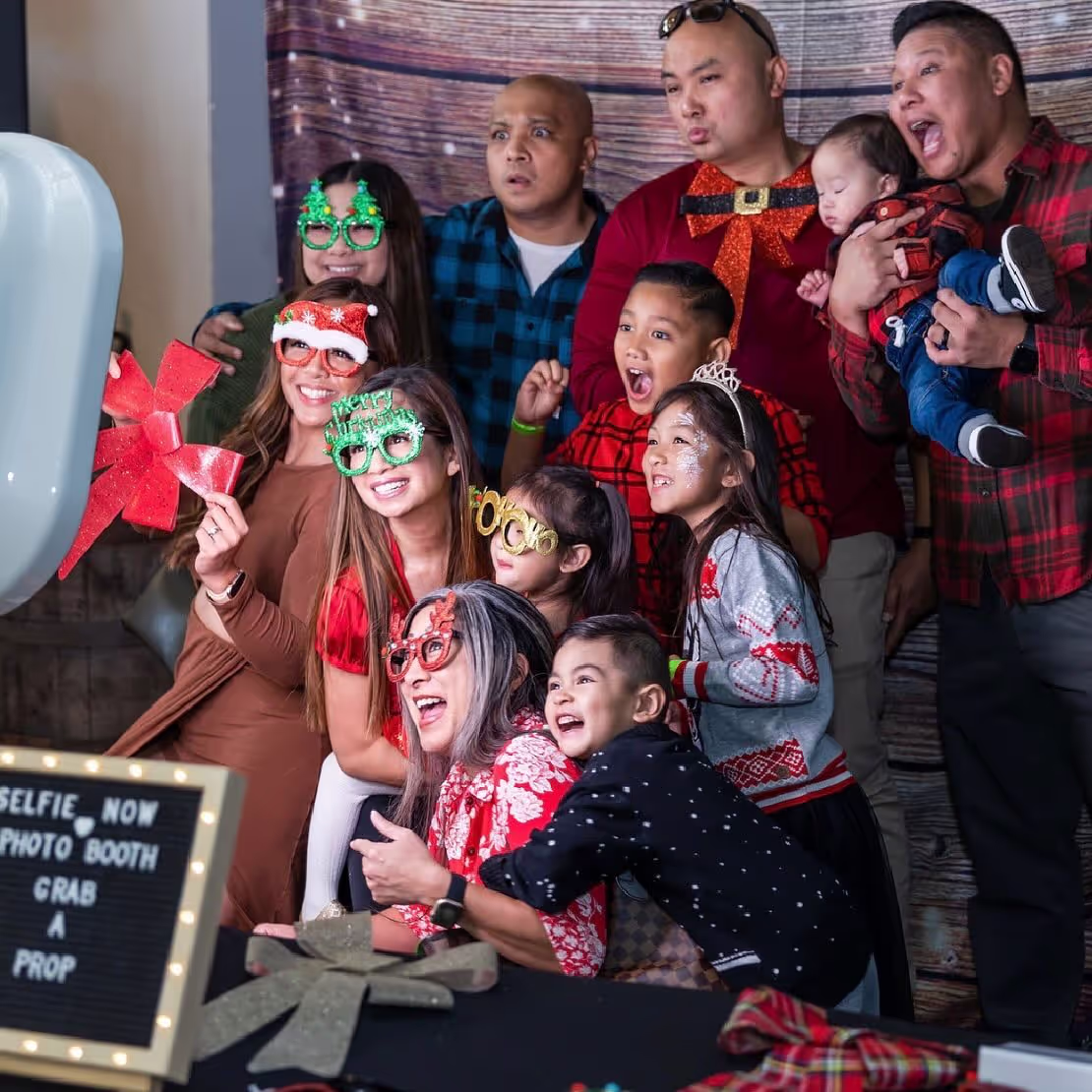 Group of adults and children wearing festive glasses and holiday attire posing excitedly in a photo booth.