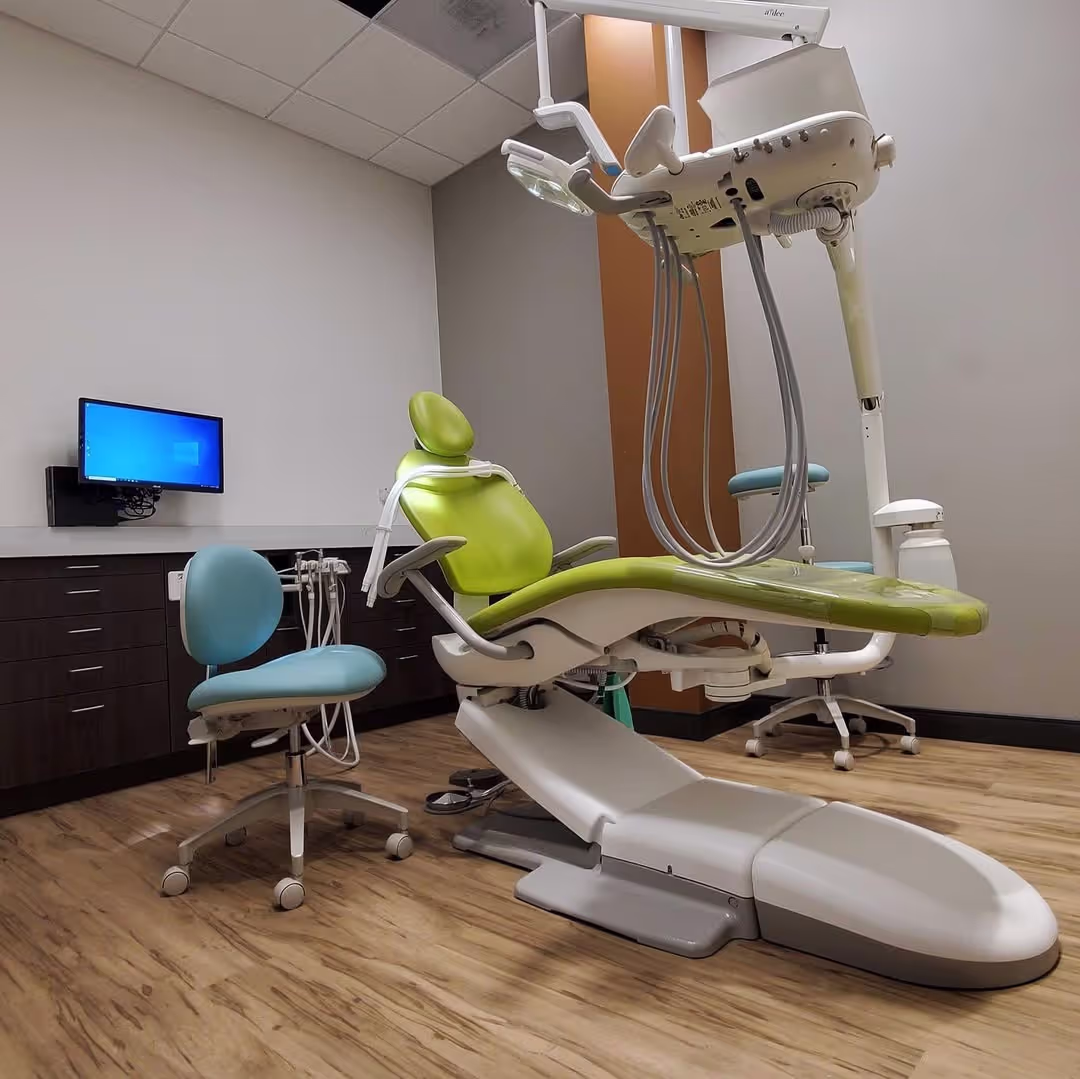 Modern dental examination room with green dentist chair, blue assistant stool, dental equipment, and a wall-mounted computer monitor.