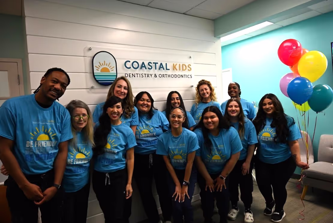 Group of smiling dental staff wearing matching blue 'Be Friendly' t-shirts posing inside Coastal Kids Dentistry & Orthodontics office with colorful balloons.