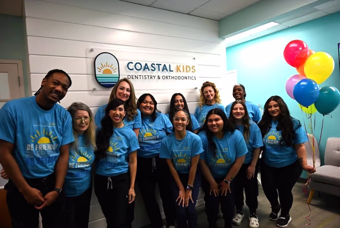 A group of smiling staff members wearing blue 'Be Friendly' Coastal Kids Dentistry T-shirts standing in a dental office lobby with colorful balloons.