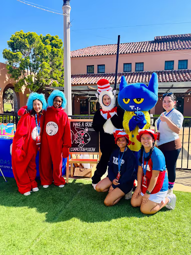 Group of six people outdoors, three in costumes including Thing 2, the Cat in the Hat, and Pete the Cat, posing in front of a building on green grass.