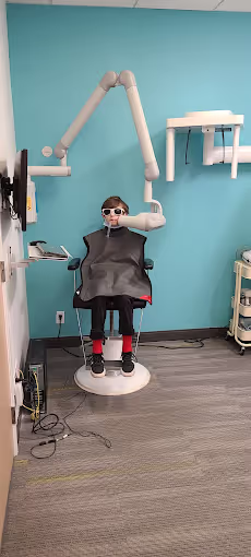 Child wearing protective eyewear and lead apron sitting in dental X-ray chair with X-ray machine positioned nearby.