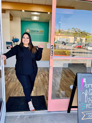 Smiling woman in black outfit standing in a doorway of a store with a pink frame door and a Coastal MD sign inside.