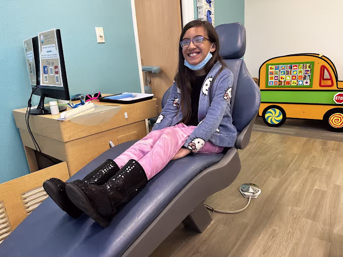 Smiling girl wearing glasses and pink pants sitting on a blue dental chair in a pediatric dental office.