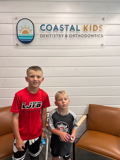 Two boys standing in front of a Coastal Kids Dentistry & Orthodontics sign on a white paneled wall, with brown chairs nearby.