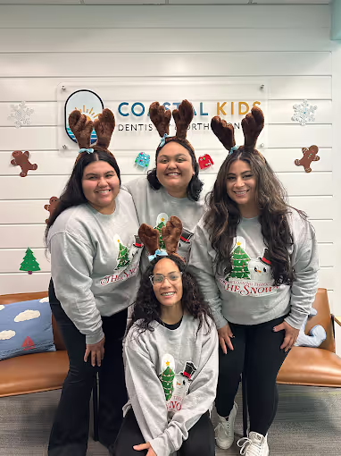 Four women wearing reindeer antler headbands and matching gray Christmas sweaters posing in front of a Coastal Kids Dentistry sign with festive holiday decorations.