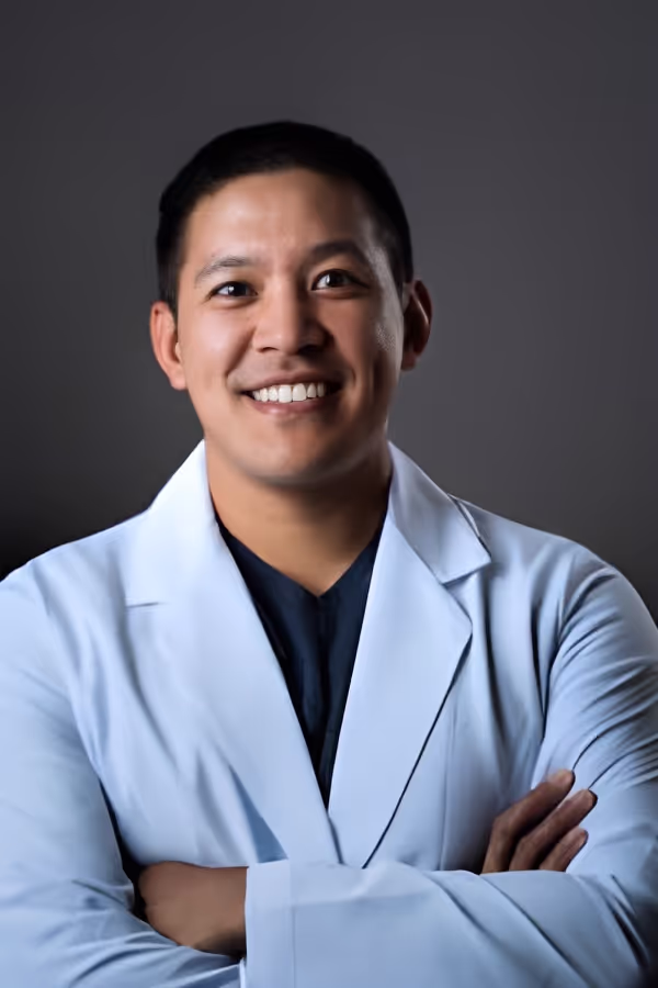 Smiling male doctor in a white lab coat with arms crossed against a dark background.