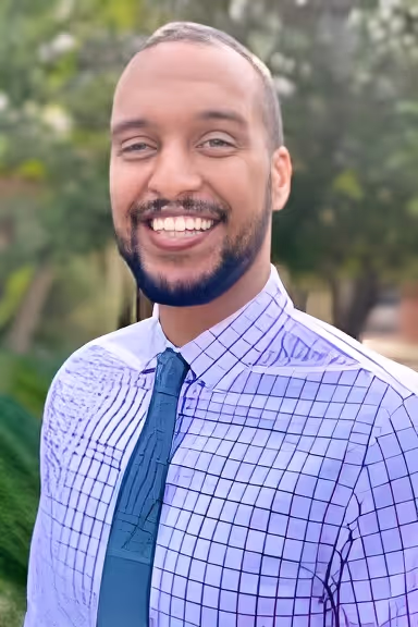 Man with beard and short hair smiling outdoors, wearing a checkered shirt and dark tie.