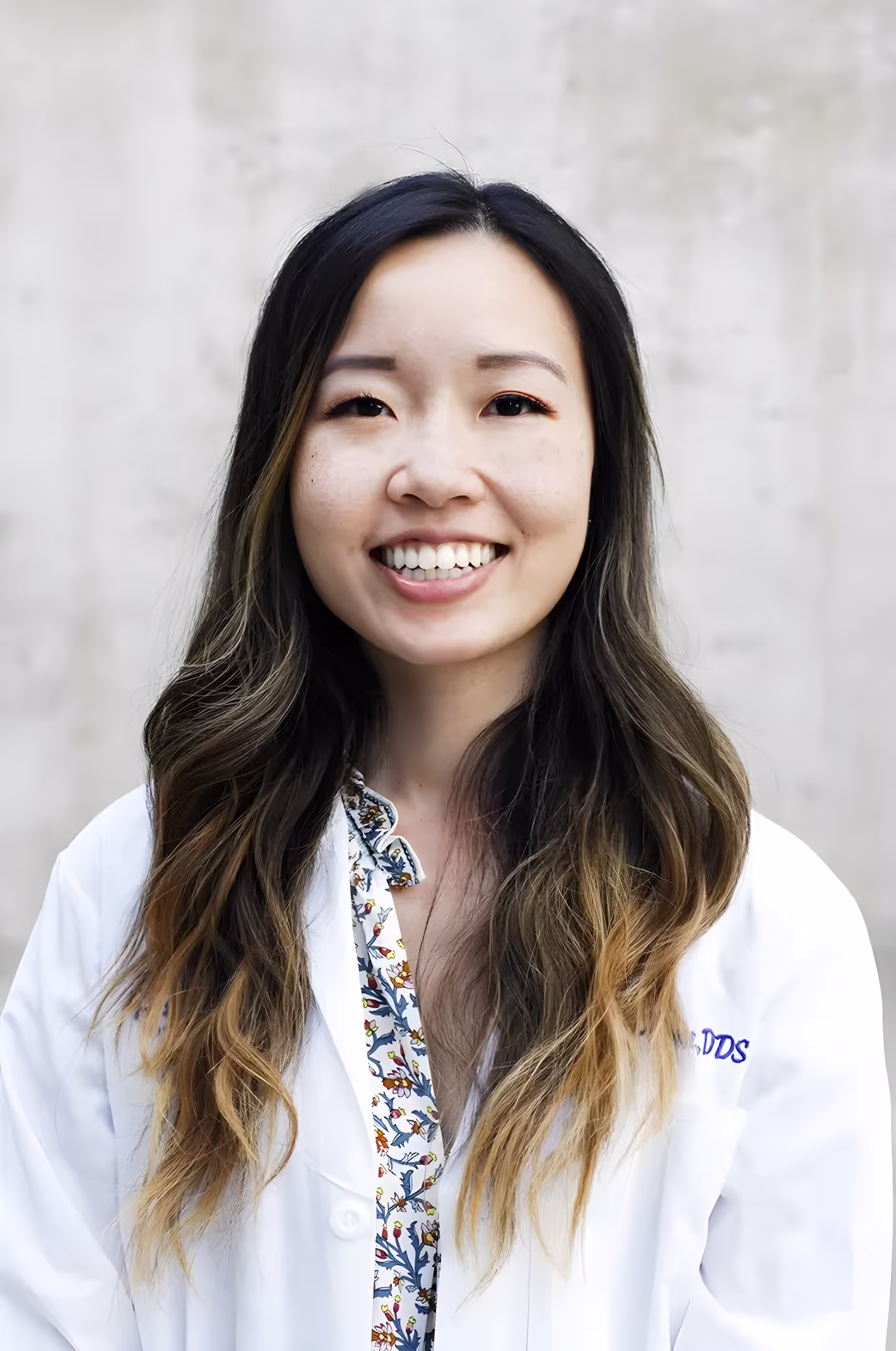Smiling Asian woman with long ombre hair wearing a white medical coat and floral blouse against a plain background.