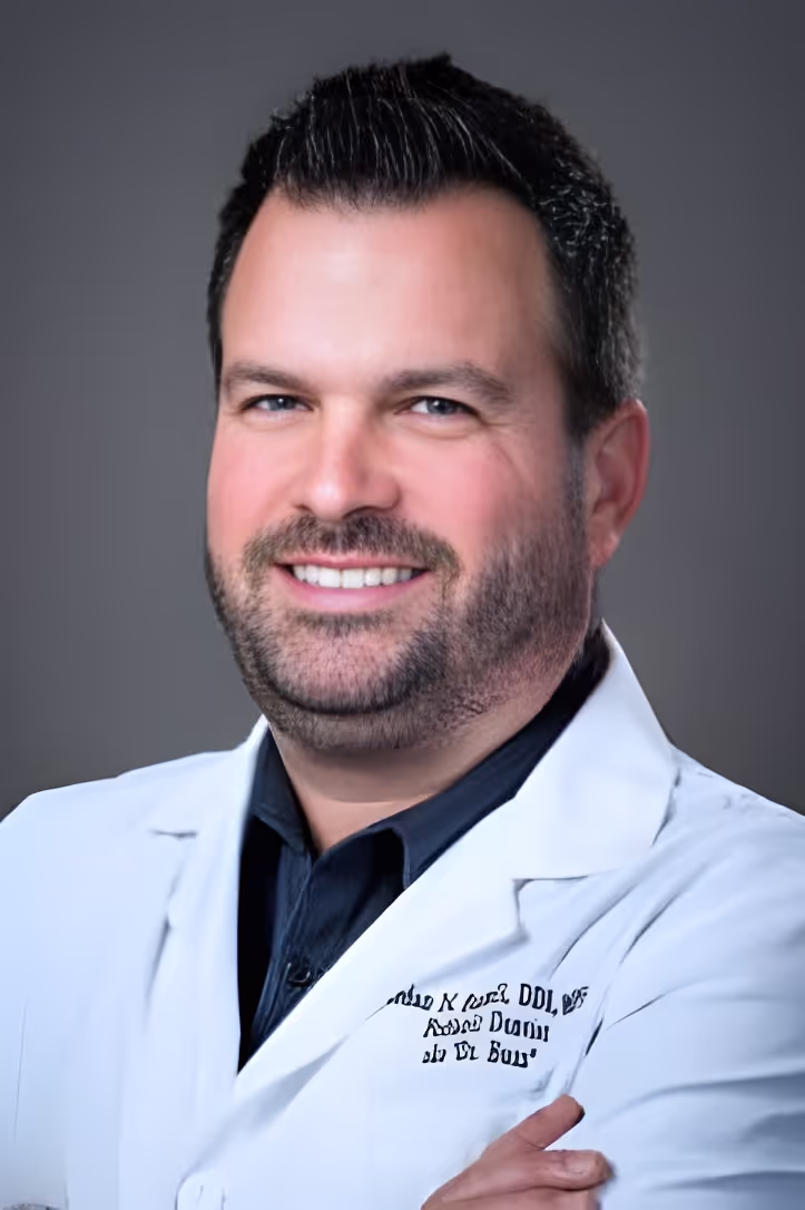 Smiling man with short dark hair and beard wearing a white medical coat over a dark shirt, arms crossed.