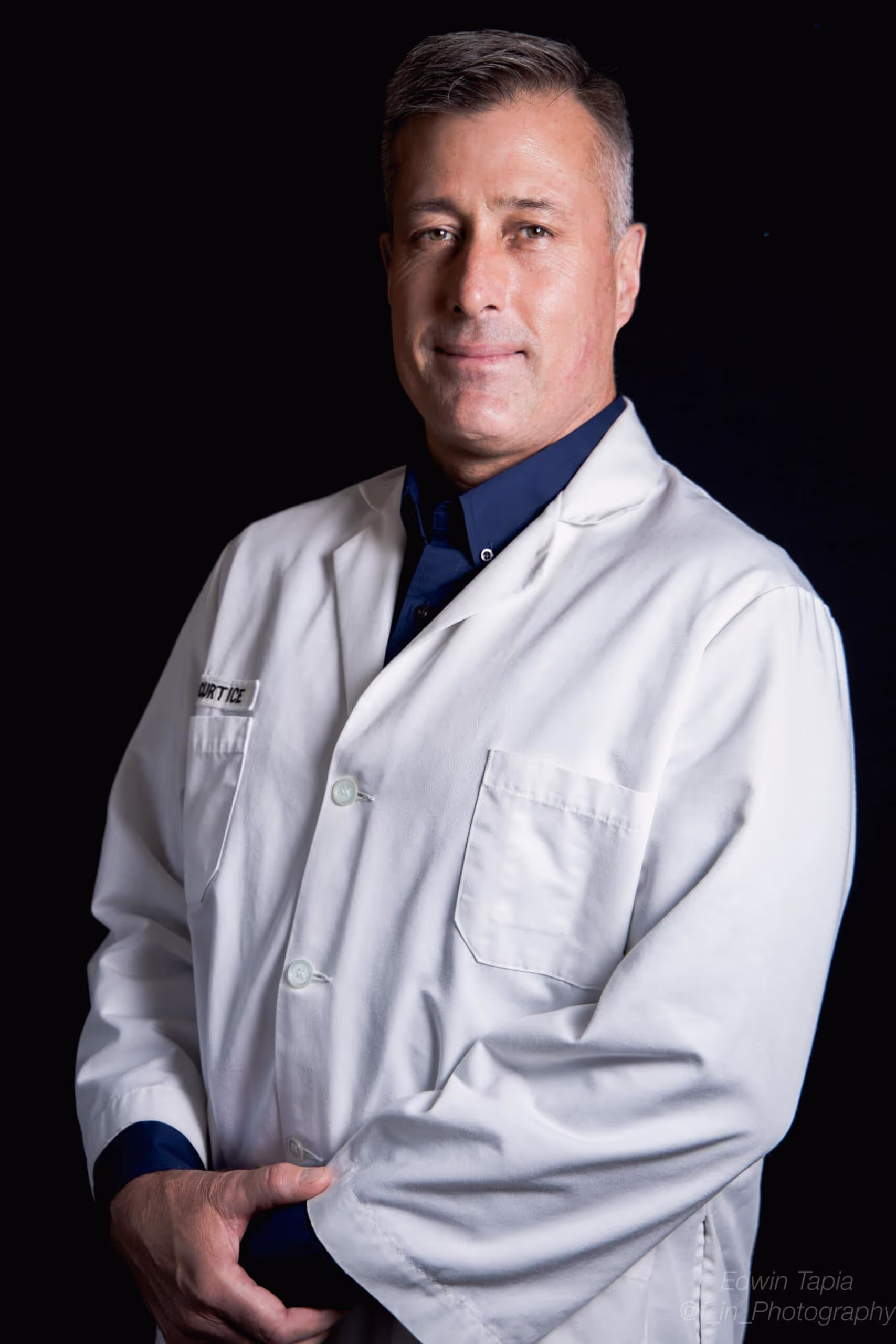Man wearing a white lab coat over a dark shirt, standing against a black background.