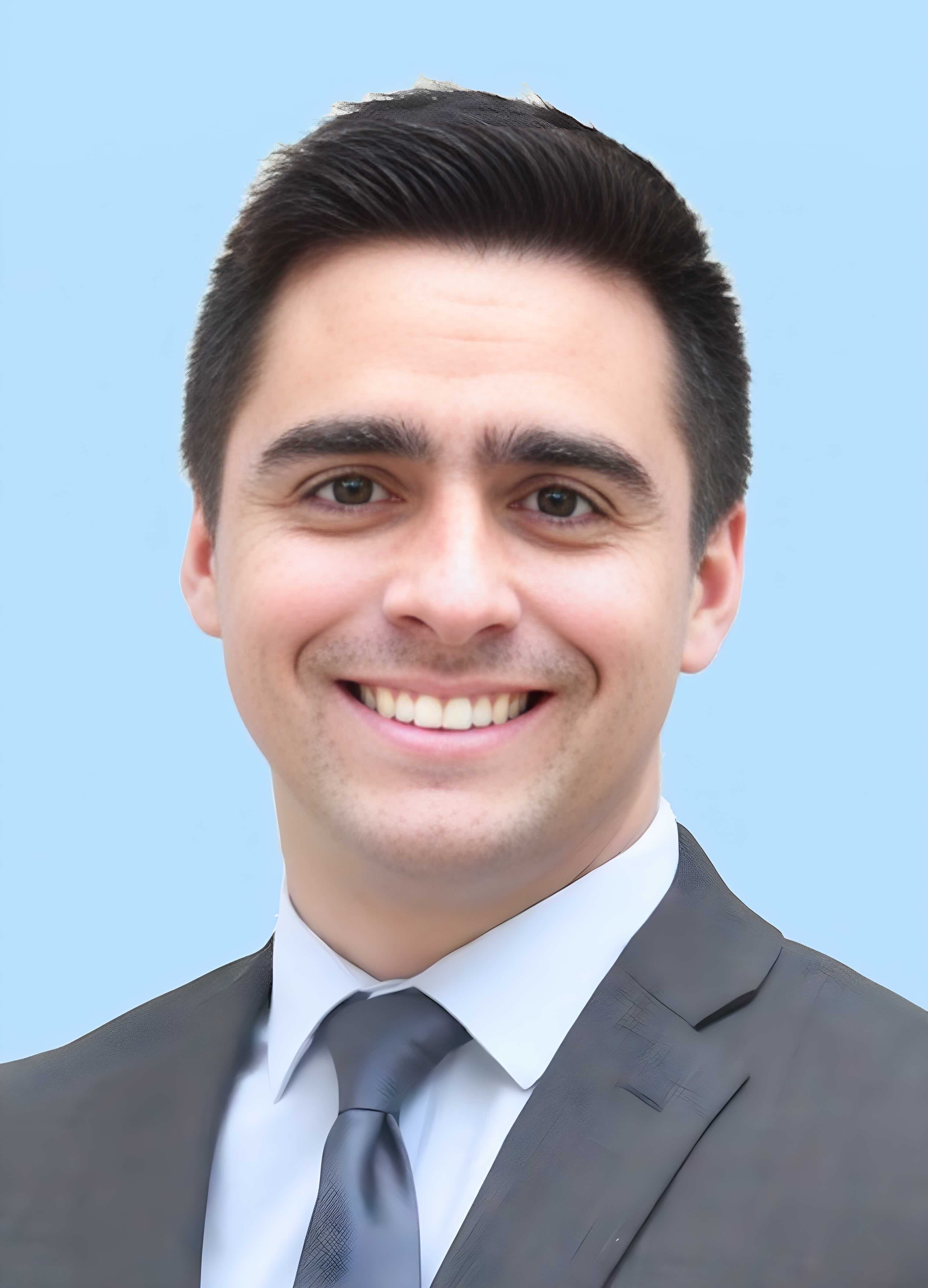 Smiling man with dark hair in a suit and tie against a light blue background.