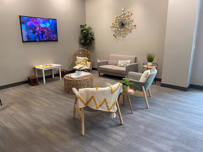 Modern living room with a gray sofa, two armchairs, a round wicker coffee table, a wall-mounted TV, wall art, and plants.