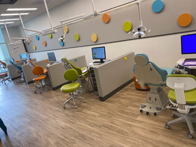 Modern dental clinic room with colorful dental chairs in green, orange, and blue, each accompanied by computer monitors and overhead dental lights.