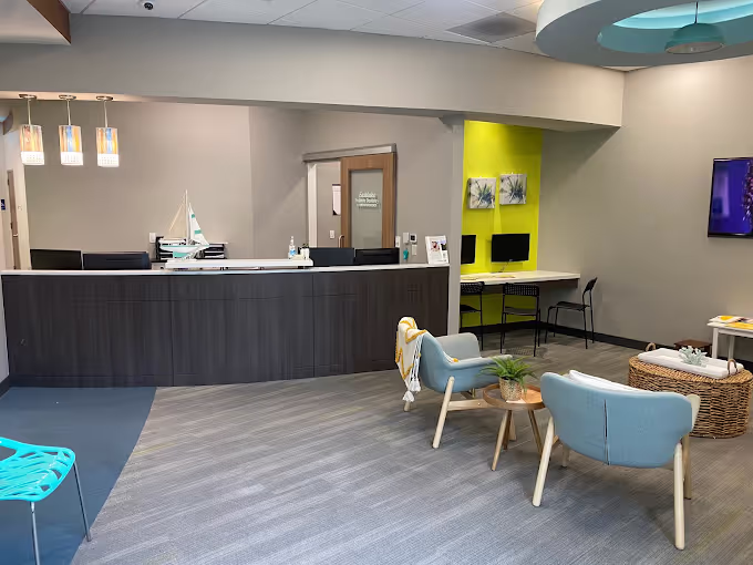 Modern office waiting area with a dark wooden reception desk, two light blue armchairs around a small wooden table with a plant, and a workspace with two computers on a lime green accent wall.