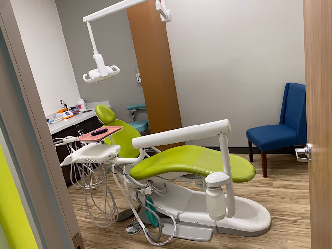 Dental clinic room with a green dental chair, dental equipment, and blue visitor chair on wood flooring.