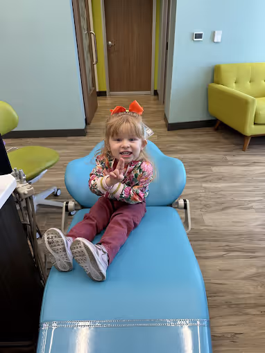 Smiling young girl with an orange bow on a blue dental chair in a clinic room.