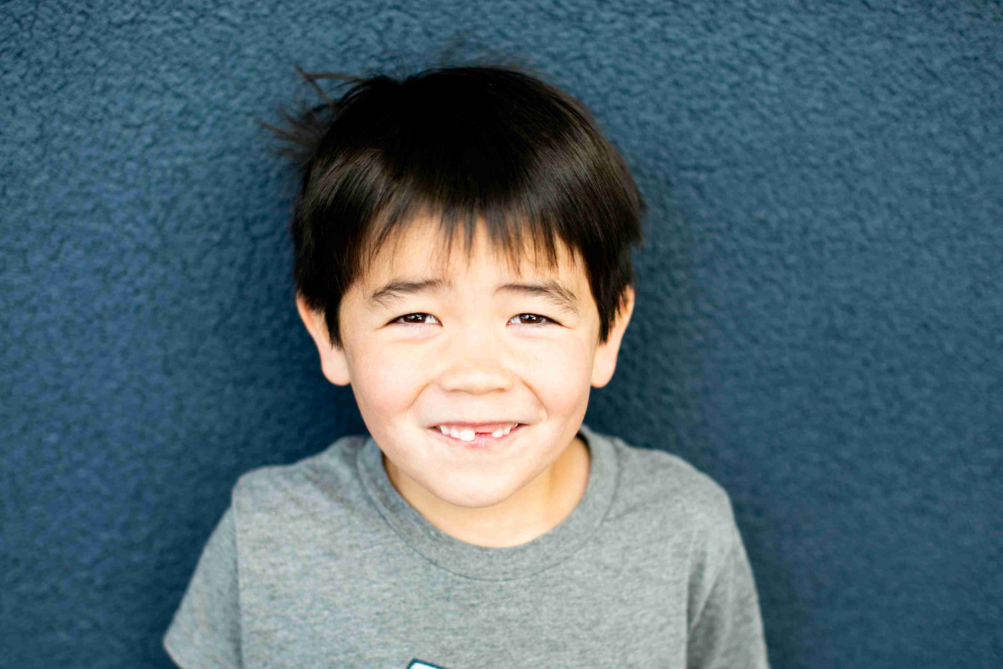 Smiling young boy with dark hair wearing a gray t-shirt against a blue textured wall.