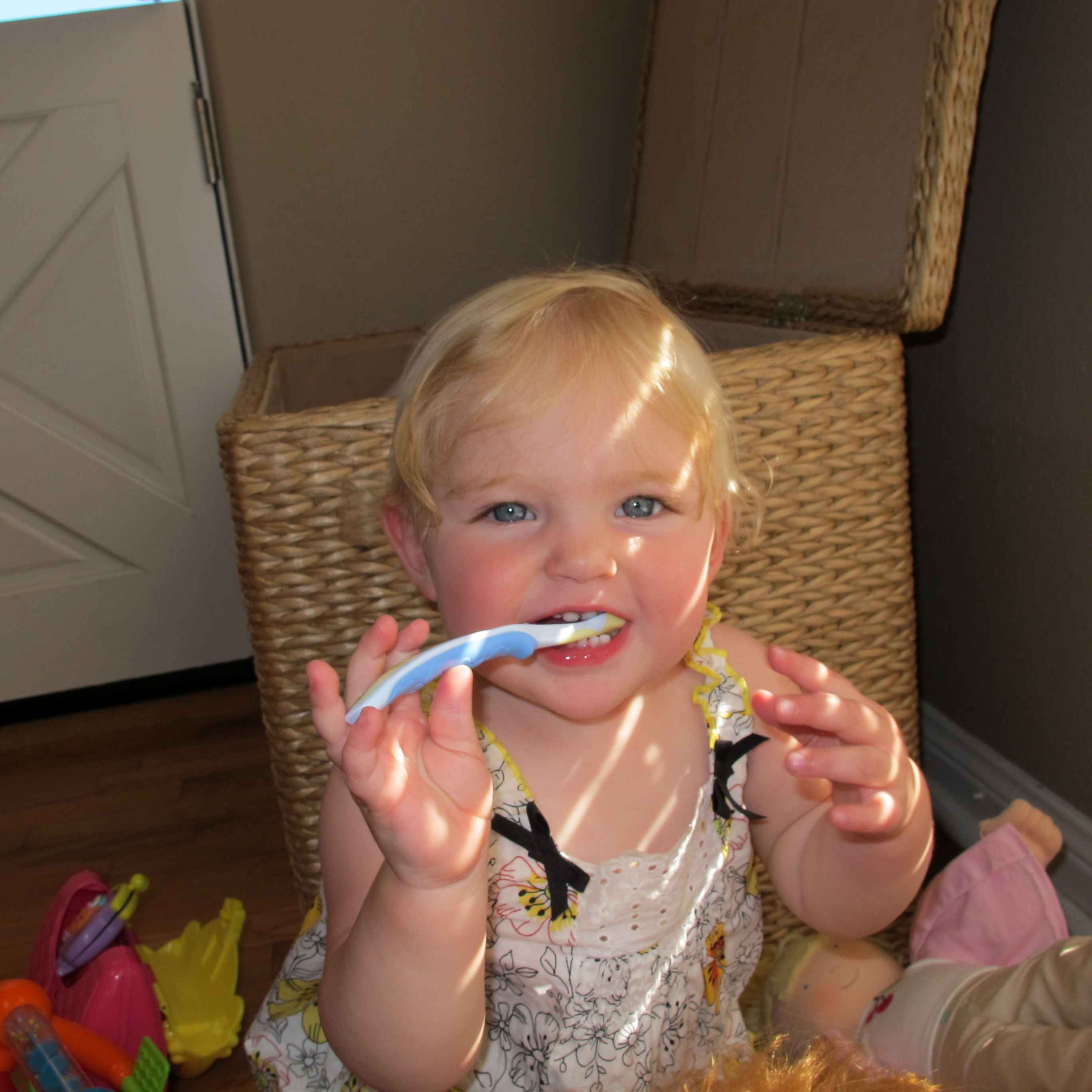 A toddler with blonde hair and blue eyes brushing their teeth with a blue toothbrush while sitting in front of a wicker basket.