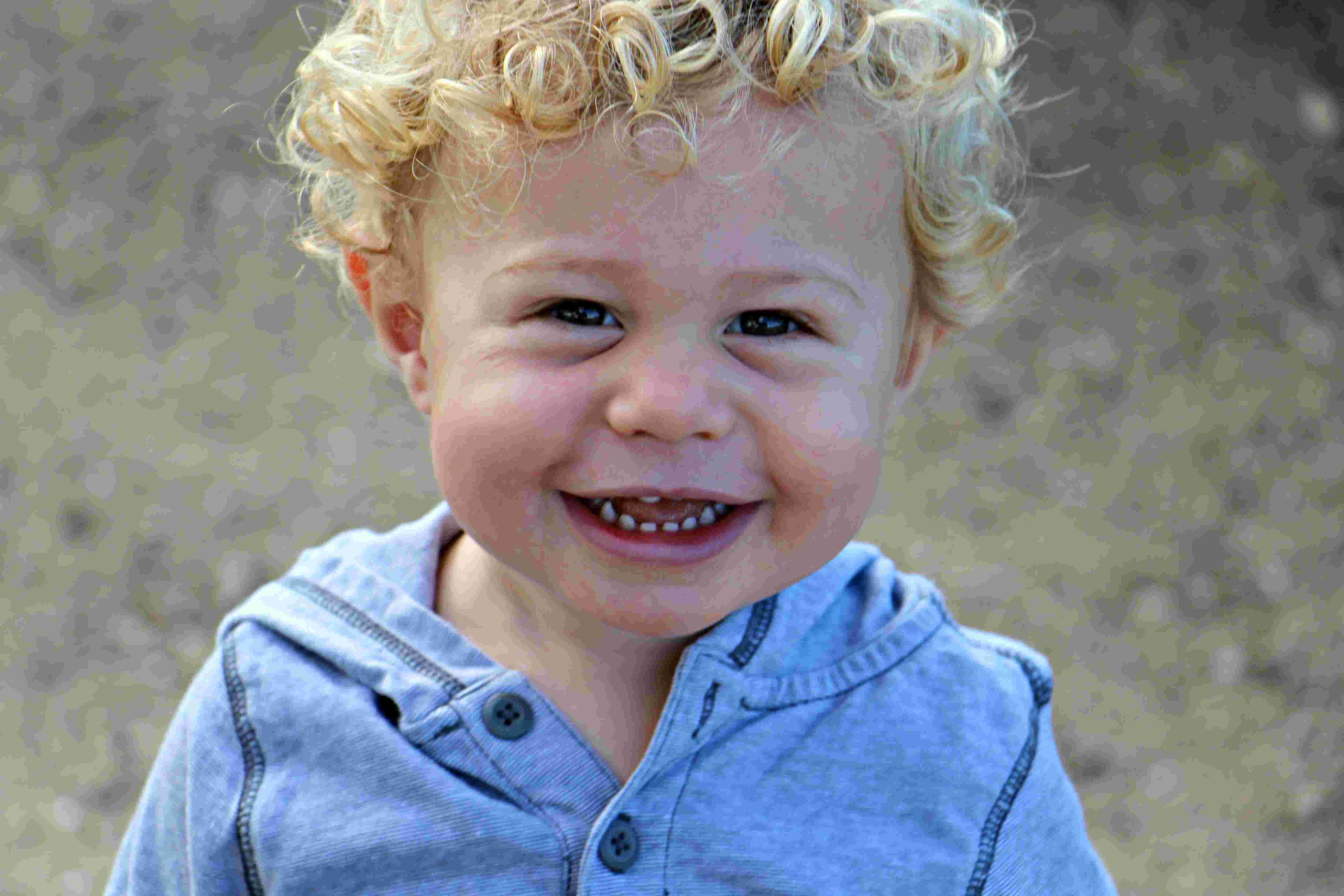 Smiling toddler with curly blonde hair wearing a blue button-up shirt outdoors.
