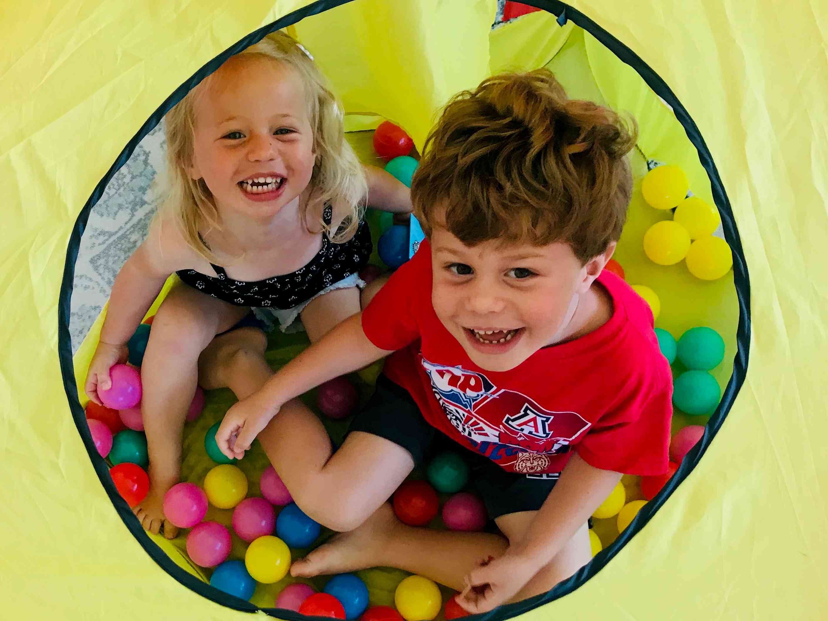 Two smiling children sitting inside a yellow play tent filled with colorful plastic balls.