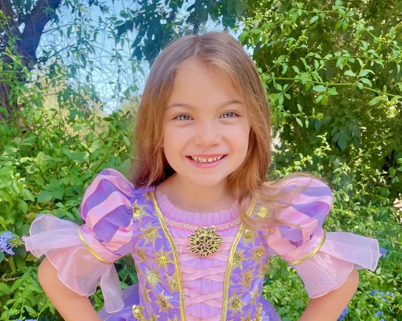 Smiling young girl in a purple and pink princess dress with gold star decorations in a green garden.