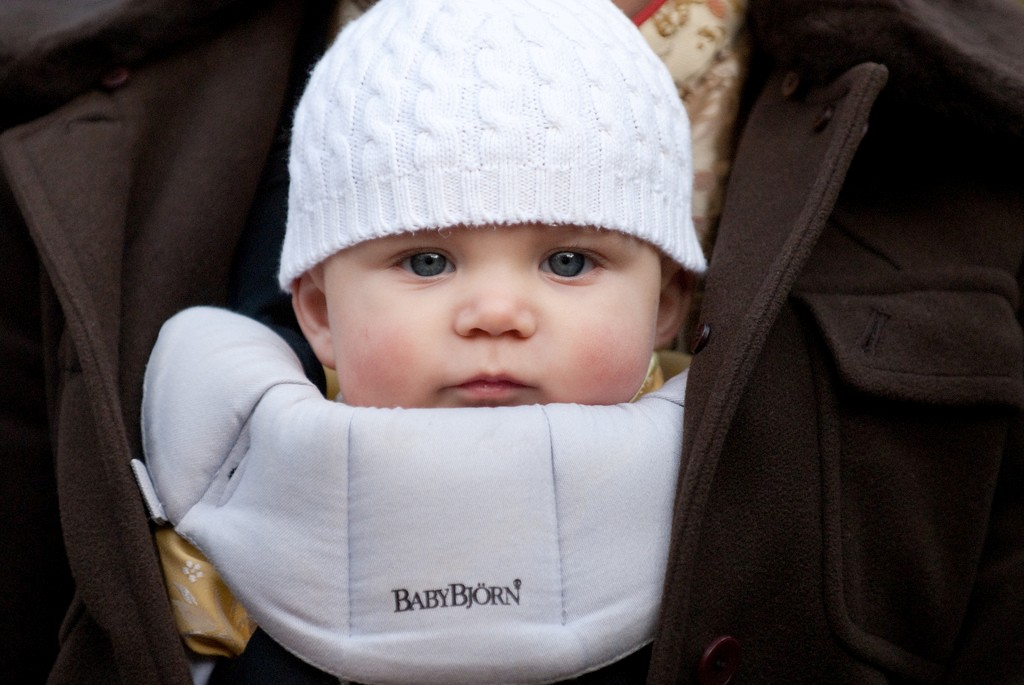 Baby with blue eyes wearing a white knit hat and gray BabyBjörn carrier, nestled inside a brown coat.