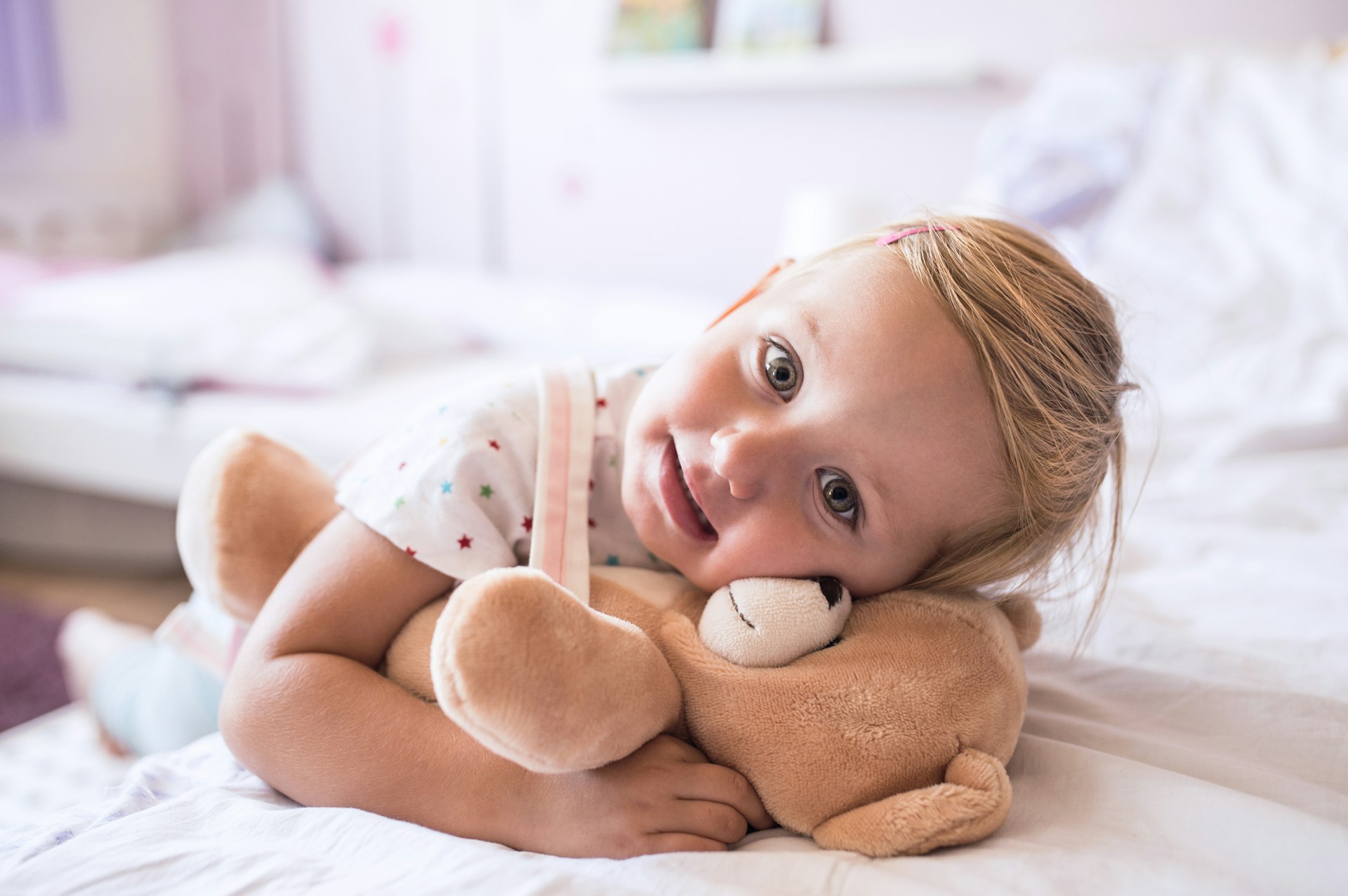 Young child lying on a bed hugging a plush teddy bear and smiling.