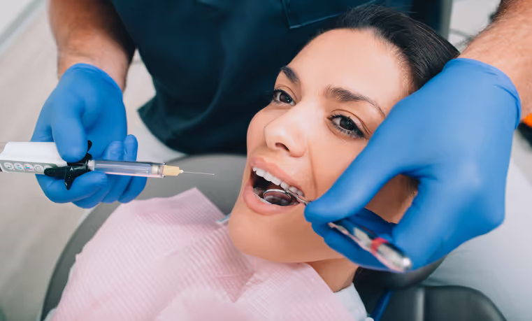 Woman in a dental chair receiving an injection from a dentist wearing blue gloves.
