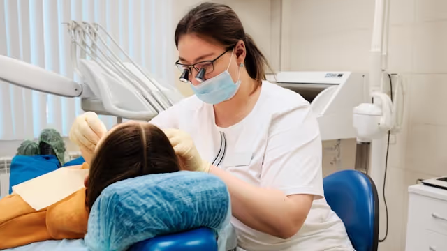 Female dentist wearing mask and loupes examining patient's teeth in a dental clinic.