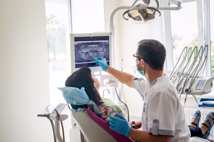 Dentist wearing gloves and a mask pointing at a dental X-ray on a monitor while discussing it with a female patient seated in a dental chair.