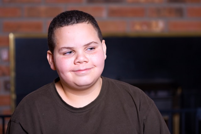Smiling young boy with short hair wearing a dark shirt sitting indoors in front of a brick wall and fireplace.