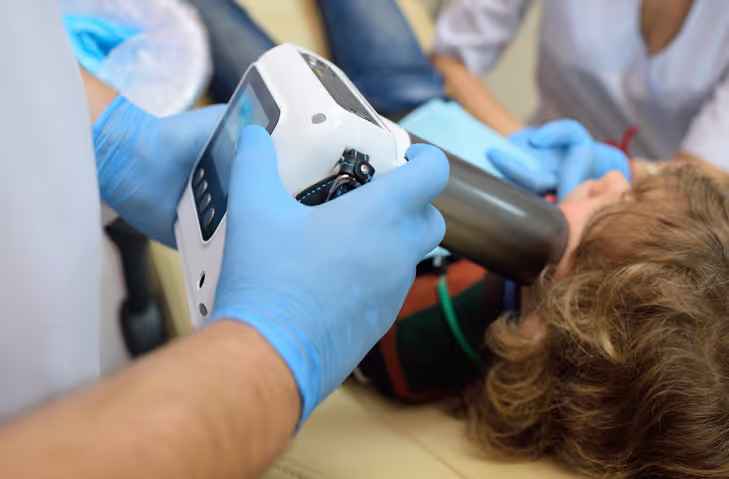 Medical professional wearing blue gloves operating a handheld X-ray device on a child's mouth while the child lies down.