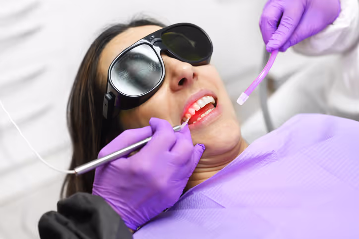 Patient wearing protective glasses receiving laser dental treatment from a dentist wearing purple gloves.