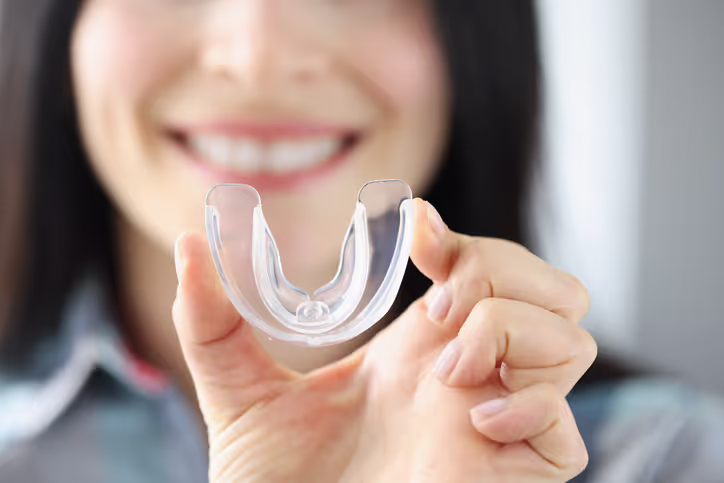 Person holding a clear dental mouthguard between thumb and forefinger in front of a smiling face.