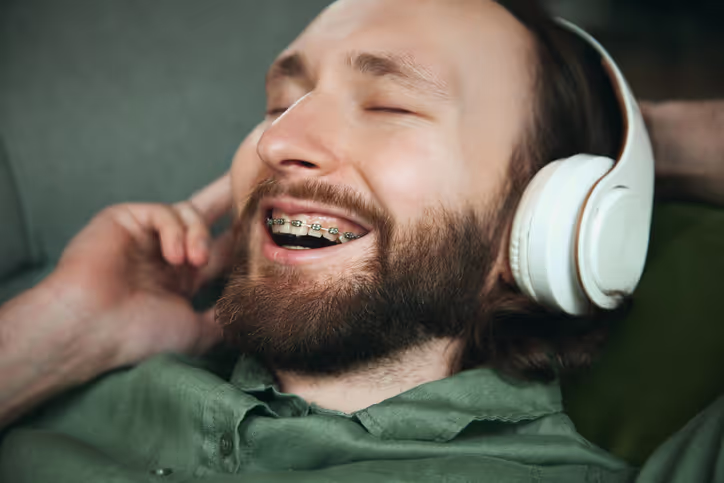 Bearded man with braces smiling with eyes closed while wearing white over-ear headphones.