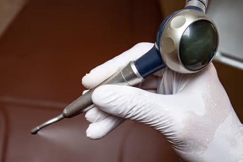 Hand wearing a white glove holding a dental prophy jet polisher tool against a brown background.