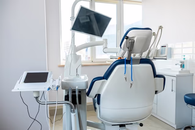 Empty dental chair in a bright clinic room with attached screen and dental equipment nearby.