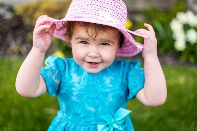 Smiling toddler in a blue dress and pink sun hat adjusting the hat outdoors with green grass and flowers in the background.